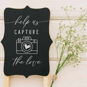 May include: Black chalkboard sign with white text that reads "Help us capture the love" with a white outline of a camera with a heart inside. The sign is propped up on a wooden stand and there are white flowers in the background.