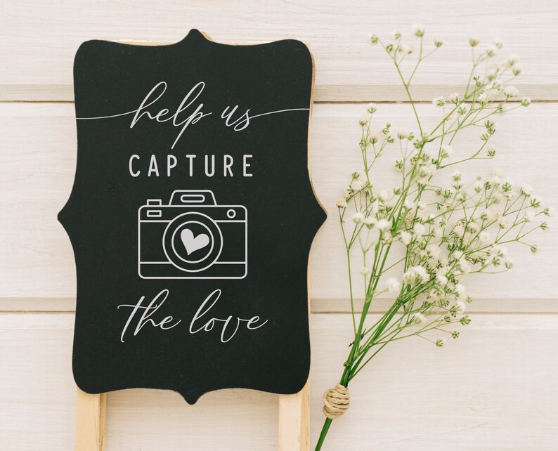 May include: Black chalkboard sign with white text that reads "Help us capture the love" with a white outline of a camera with a heart inside. The sign is propped up on a wooden stand and there are white flowers in the background.