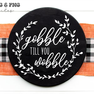 May include: A circular black wooden sign featuring white text that reads "gobble TILL YOU wobble" centered within a white leafy vine wreath design. The sign is positioned over a horizontal orange burlap ribbon and a black and white buffalo check ribbon.