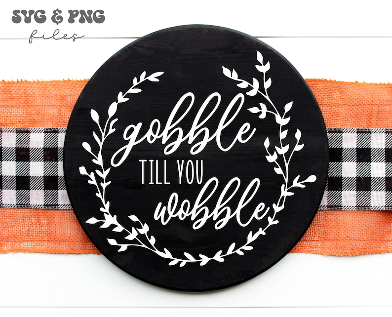 May include: A circular black wooden sign featuring white text that reads "gobble TILL YOU wobble" centered within a white leafy vine wreath design. The sign is positioned over a horizontal orange burlap ribbon and a black and white buffalo check ribbon.