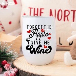 May include: White wine tumbler with black text that reads "Forget the Apple Give me Wine". The text is surrounded by red hearts. The tumbler is sitting on a wooden surface.