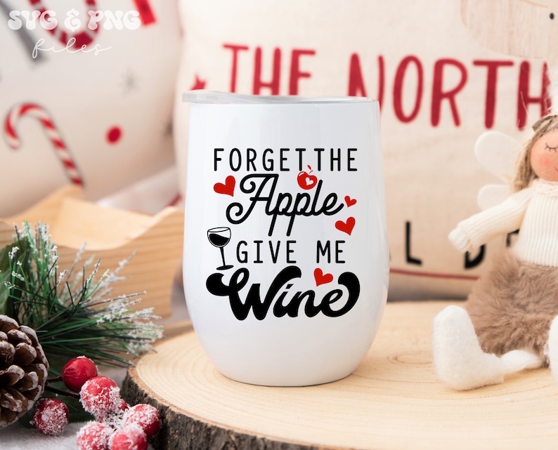 May include: White wine tumbler with black text that reads "Forget the Apple Give me Wine". The text is surrounded by red hearts. The tumbler is sitting on a wooden surface.