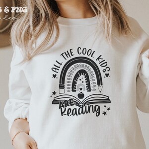 May include: A white crewneck sweatshirt featuring a black graphic print of an open book with an apple in the center, topped by a rainbow made of pencils and paperclips, with the text ALL THE COOL KIDS ARE READING surrounding the design.