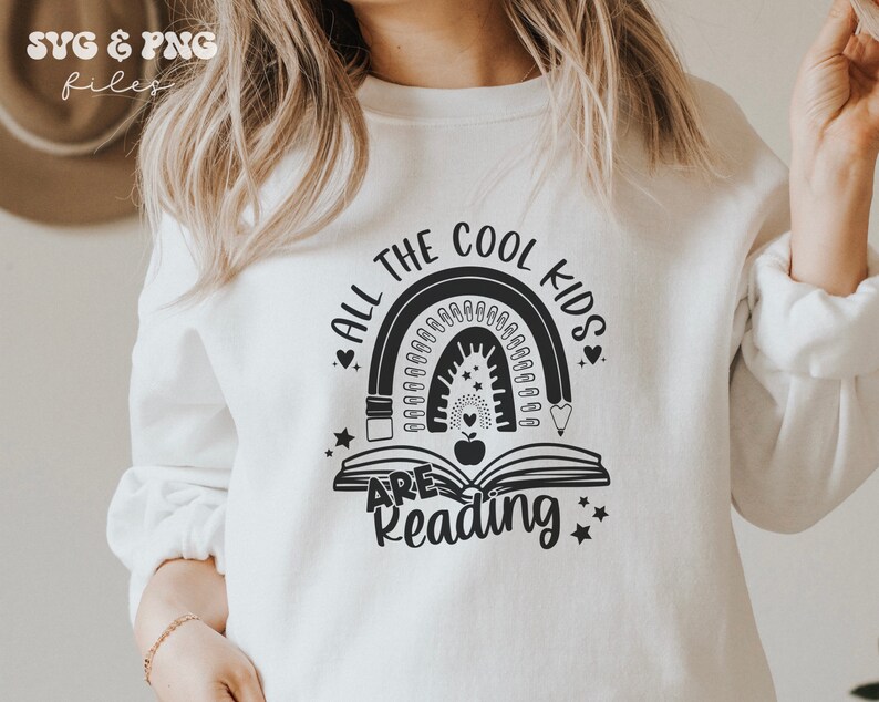 May include: A white crewneck sweatshirt featuring a black graphic print of an open book with an apple in the center, topped by a rainbow made of pencils and paperclips, with the text ALL THE COOL KIDS ARE READING surrounding the design.
