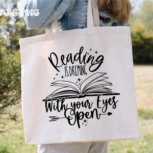 May include: A white canvas tote bag featuring a black graphic design of an open book with the text Reading is dreaming with your eyes open, accented by small stars and an arrow. The top left corner displays the text SVG & PNG files.