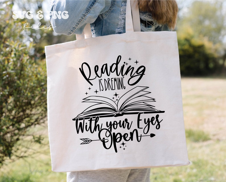 May include: A white canvas tote bag featuring a black graphic design of an open book with the text Reading is dreaming with your eyes open, accented by small stars and an arrow. The top left corner displays the text SVG & PNG files.
