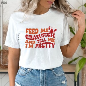 May include: White t-shirt with the text "Feed me crawfish and tell me I'm pretty" in a retro font. A red crawfish illustration is above the text.
