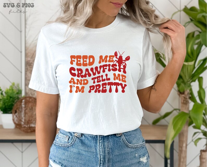 May include: White t-shirt with the text "Feed me crawfish and tell me I'm pretty" in a retro font. A red crawfish illustration is above the text.