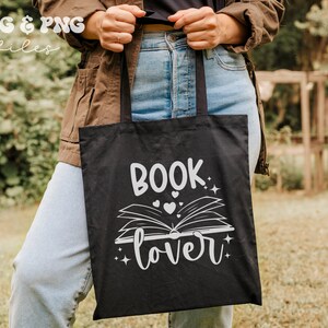 May include: A black tote bag featuring a white graphic design of an open book with the text "BOOK lover" above and below the illustration, accented with small hearts and star shapes. The bag has two black fabric handles and a rectangular shape.