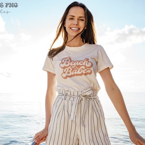 May include: A woman wearing a white t-shirt with the text "Beach Babe" in pink script with palm trees. She is standing in front of a blue body of water and a cloudy sky.