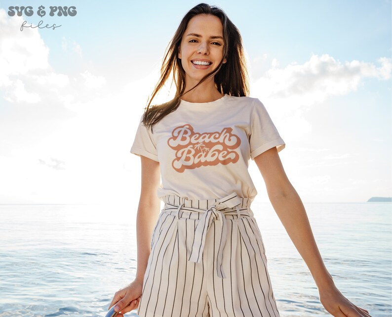 May include: A woman wearing a white t-shirt with the text "Beach Babe" in pink script with palm trees. She is standing in front of a blue body of water and a cloudy sky.