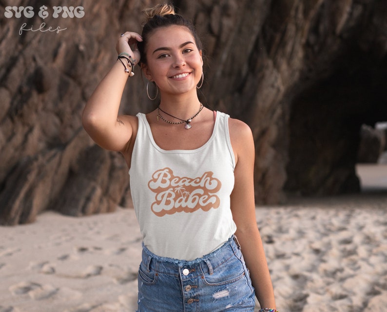 May include: A white tank top with a brown retro-style graphic that says "Beach Babe" with a palm tree icon. The model is wearing the tank top and smiling at the camera.