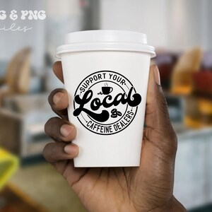 May include: A white to-go coffee cup with a black circle design that says "Support Your Local Caffeine Dealers" with a stylized coffee cup and coffee beans inside the circle.