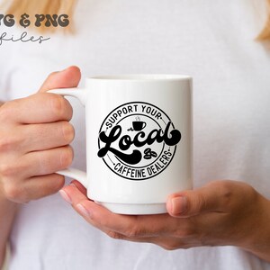 May include: White ceramic mug with a black circle design that says "Support Your Local Caffeine Dealers" with a coffee cup icon.