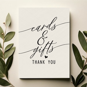 May include: A white card with black calligraphy that reads "cards & gifts thank you". The card is surrounded by green eucalyptus branches.