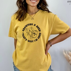 May include: A yellow t-shirt with a black graphic of a crawfish holding a can of beer. The text on the shirt reads "Crawfish & Beer... That's Why I'm Here."