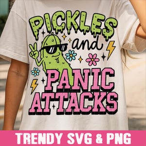 Könnte beinhalten: Cremefarbenes T-Shirt mit dem Aufdruck "Pickles and Panic Attacks". Das Design zeigt eine Cartoon-Gurke mit Sonnenbrille und Peace-Zeichen, sowie Blumen und Blitze.