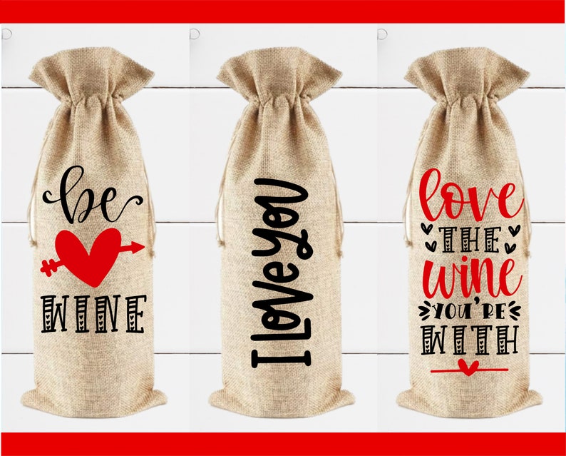 May include: Three burlap wine bottle bags with different Valentine's Day themed sayings. The first bag says "be wine" with a red heart and arrow. The second bag says "I love you". The third bag says "love the wine you're with".