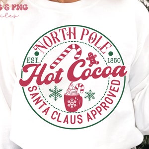 May include: A white sweatshirt with a green circle design that says "North Pole Est. 1850 Hot Cocoa Santa Claus Approved" with a red candy cane, gingerbread man, and a cup of hot chocolate with whipped cream and a candy cane.