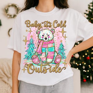 May include: White t-shirt with a bear wearing a colourful scarf and mittens. The design includes Christmas trees and the gold text "Baby it's Cold Outside". A festive holiday design, perfect for the winter season.