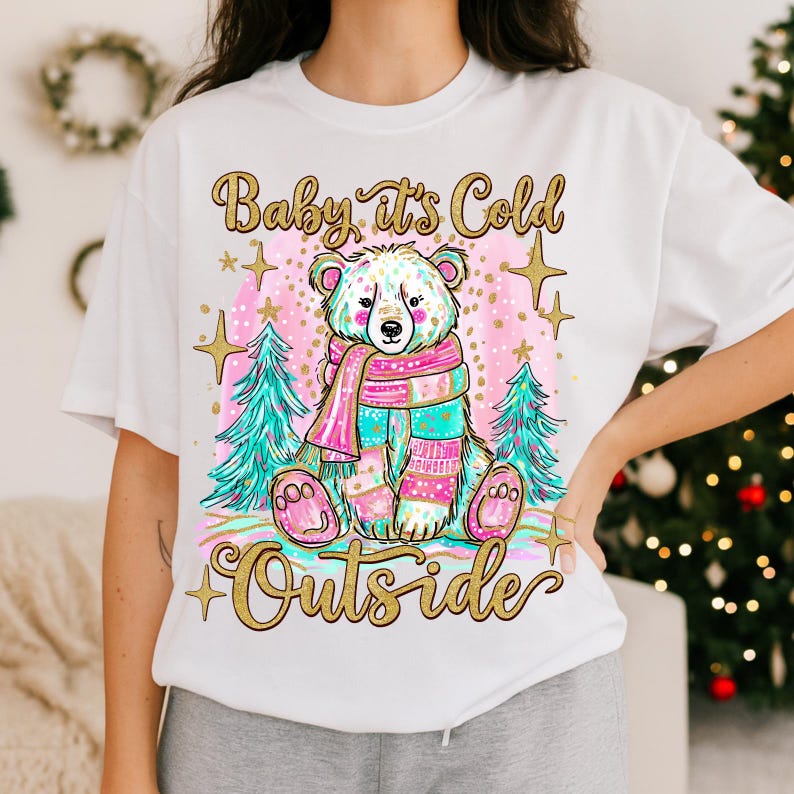 May include: White t-shirt with a bear wearing a colourful scarf and mittens. The design includes Christmas trees and the gold text "Baby it's Cold Outside". A festive holiday design, perfect for the winter season.