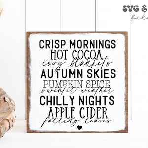 May include: A square wooden sign with a white center and dark wood frame. The text reads: CRISP MORNINGS, HOT COCOA, cozy blankets, AUTUMN SKIES, PUMPKIN SPICE, sweater weather, CHILLY NIGHTS, APPLE CIDER, falling leaves, and a small black heart.