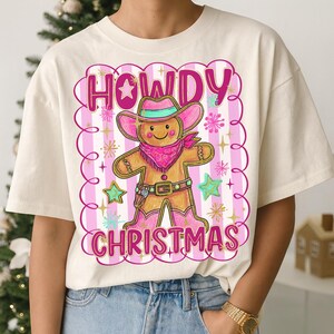 May include: Cream-coloured t-shirt with a gingerbread cowboy design. The graphic features the words "HOWDY CHRISTMAS" in pink, a gingerbread man wearing a pink cowboy hat and boots, and star accents.
