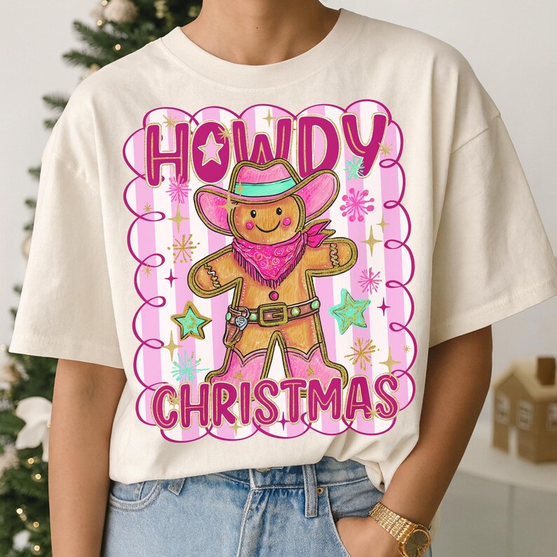 May include: Cream-coloured t-shirt with a gingerbread cowboy design. The graphic features the words "HOWDY CHRISTMAS" in pink, a gingerbread man wearing a pink cowboy hat and boots, and star accents.