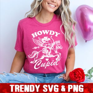 Op de afbeelding: Een roze T-shirt met een witte afbeelding van een cupido die op een paard rijdt met de tekst "Howdy Cupid".
