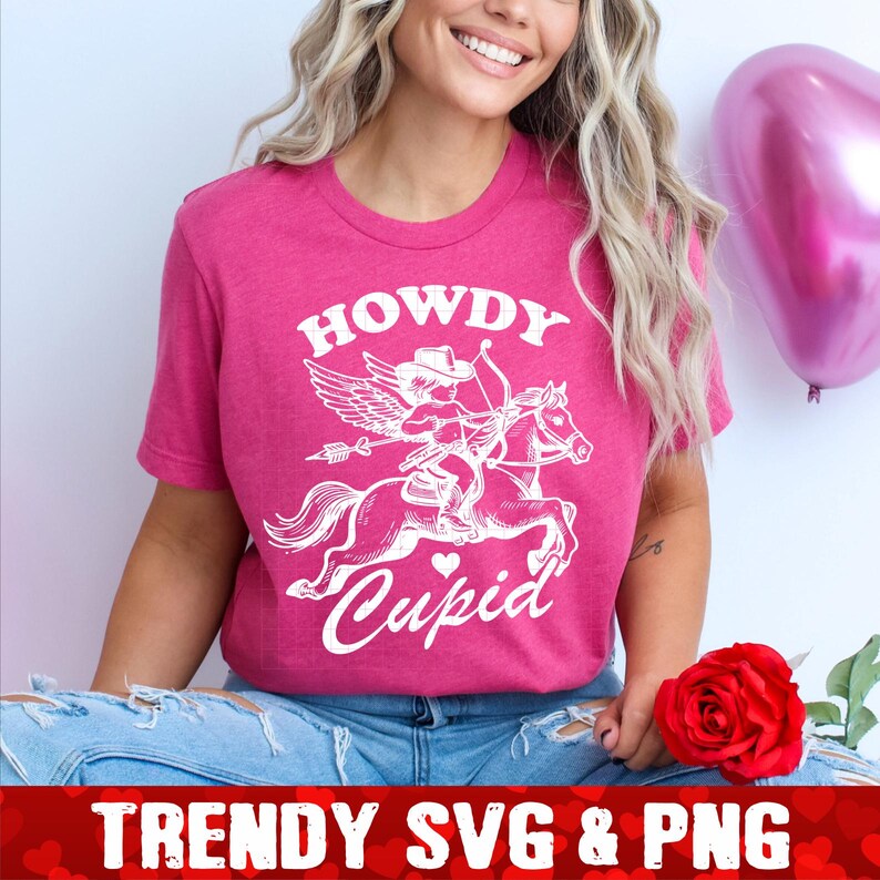 Op de afbeelding: Een roze T-shirt met een witte afbeelding van een cupido die op een paard rijdt met de tekst "Howdy Cupid".