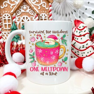 May include: White ceramic mug with a festive holiday design. The mug features the text "Surviving the Holidays, One Meltdown at a Time." A pink cup with a snowman in a hot chocolate bath is the central graphic. Gold stars and snowflakes decorate the mug.