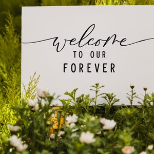 May include: White sign with black calligraphy that reads "Welcome to our Forever" surrounded by green foliage and white flowers.