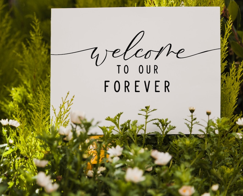 May include: White sign with black calligraphy that reads "Welcome to our Forever" surrounded by green foliage and white flowers.