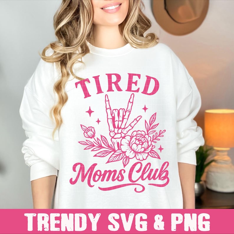 May include: A white sweatshirt with pink text that reads "TIRED MOMS CLUB". The text is in a cursive font. A pink hand sign with a skeleton hand is above the text. The hand sign is surrounded by pink flowers.
