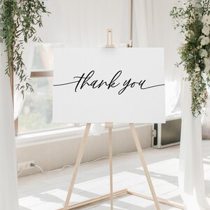 May include: A white sign with black calligraphy that reads "Thank you" on an easel in front of a white wall with greenery on either side.