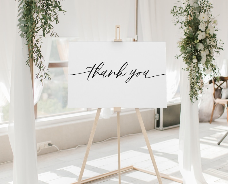 May include: A white sign with black calligraphy that reads "Thank you" on an easel in front of a white wall with greenery on either side.