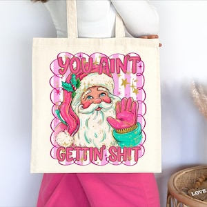 May include: A cream tote bag with a Santa Claus illustration and the text "YOU AINT GETTIN SHIT" in pink. Santa is wearing a pink hat and gloves, with a turquoise scarf and a sprig of holly.