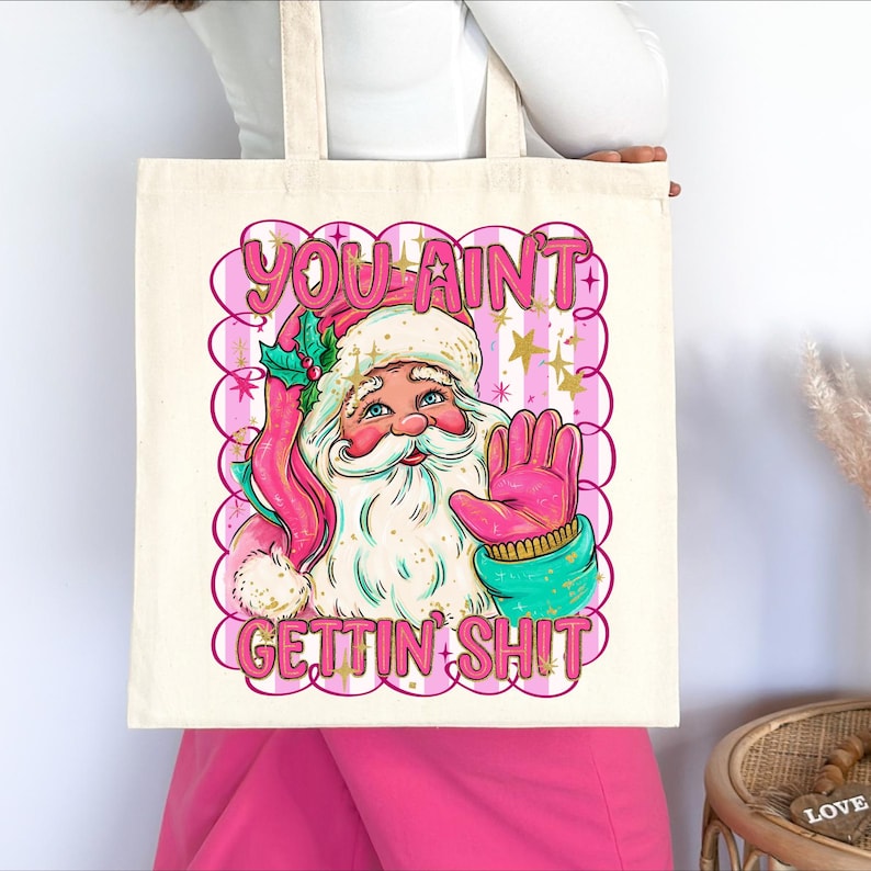 May include: A cream tote bag with a Santa Claus illustration and the text "YOU AINT GETTIN SHIT" in pink. Santa is wearing a pink hat and gloves, with a turquoise scarf and a sprig of holly.