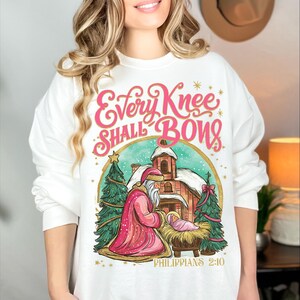 May include: White sweatshirt with a graphic featuring a Christmas scene with Santa kneeling before baby Jesus. The text "Every Knee Shall Bows" is in pink script, with "Philippians 2:10" below the image. Two green Christmas trees flank the scene.