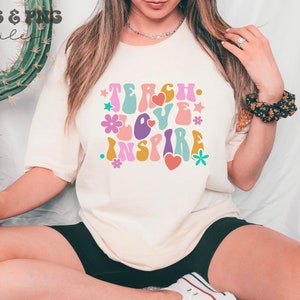 Peut inclure: Un t-shirt blanc avec un graphique de style r&eacute;tro qui dit "Teach Love Inspire" en lettres color&eacute;es et ondul&eacute;es. Le graphique comprend &eacute;galement des fleurs et des c&oelig;urs.