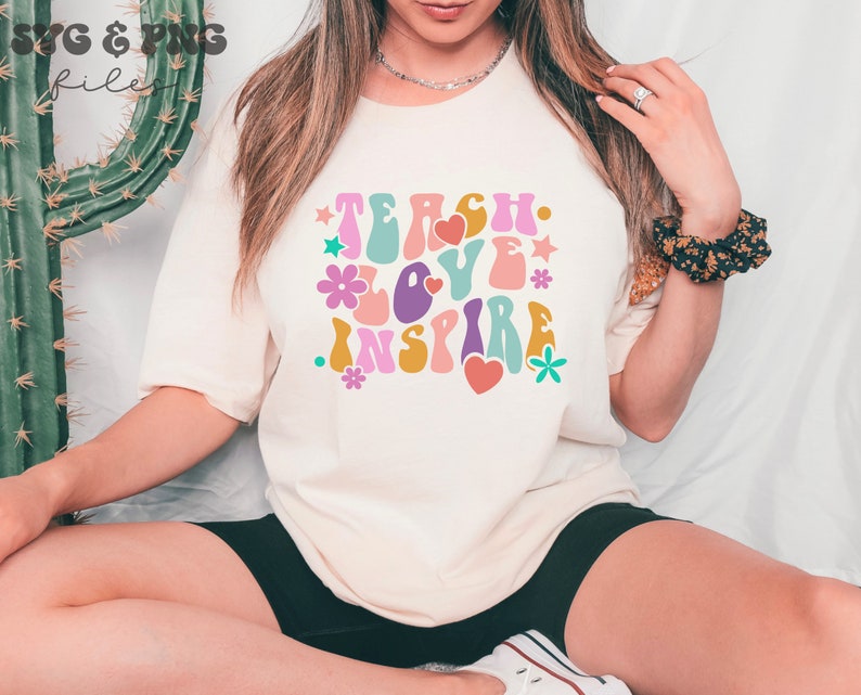 Peut inclure: Un t-shirt blanc avec un graphique de style r&eacute;tro qui dit "Teach Love Inspire" en lettres color&eacute;es et ondul&eacute;es. Le graphique comprend &eacute;galement des fleurs et des c&oelig;urs.