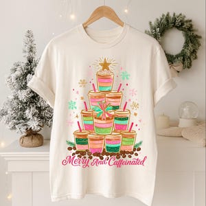May include: Off-white t-shirt with a Christmas tree design composed of layered coffee cups in pink, green, and brown. The design includes a gold star, snowflakes, and the text "Merry And Caffeinated" in pink.