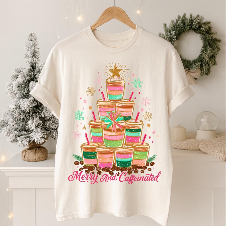 May include: Off-white t-shirt with a Christmas tree design composed of layered coffee cups in pink, green, and brown. The design includes a gold star, snowflakes, and the text "Merry And Caffeinated" in pink.