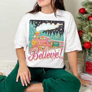 May include: White t-shirt with a colourful train illustration and the word "Believe!" in script. The train is pink and green, set against a snowy landscape and starry night. The shirt has a casual, oversized fit, suitable for everyday wear.