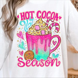 May include: White sweatshirt with a vibrant graphic. The design shows a pink and teal mug of hot chocolate with cream, marshmallows, and a candy cane. The words "HOT COCOA SEASON" are written in pink.