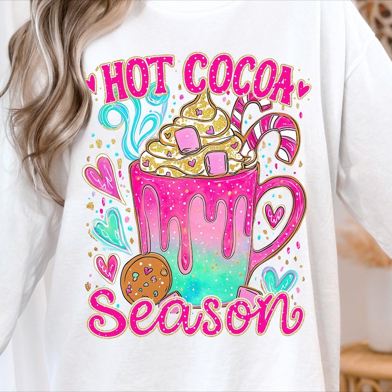 May include: White sweatshirt with a vibrant graphic. The design shows a pink and teal mug of hot chocolate with cream, marshmallows, and a candy cane. The words "HOT COCOA SEASON" are written in pink.