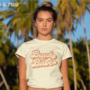May include: A white t-shirt with a retro-style graphic that says "Beach Babe" in a peach color. The graphic includes a palm tree silhouette.