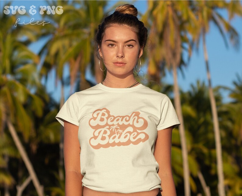 May include: A white t-shirt with a retro-style graphic that says "Beach Babe" in a peach color. The graphic includes a palm tree silhouette.