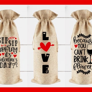 May include: Three burlap wine bottle bags with different Valentine's Day themed designs. The first bag says "Sip Sip Hooray It's Valentine's Day". The second bag says "Love" with a red heart and arrow. The third bag says "Because You Can't Drink Flowers" with a wine glass and hearts.