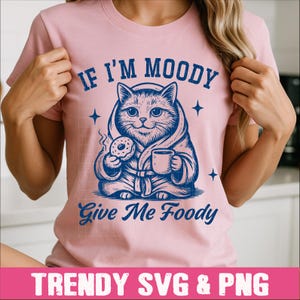 May include: Pink t-shirt with a blue illustration of a cat in a robe holding a donut and a cup of coffee. The text "If I'm Moody Give Me Foody" is printed on the shirt.